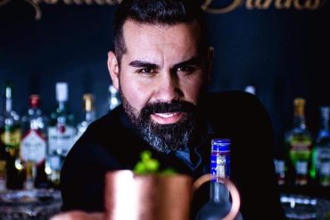 Homem sorridente misturando drink colorido em cozinha moderna, curso de bebidas Gui Rial.